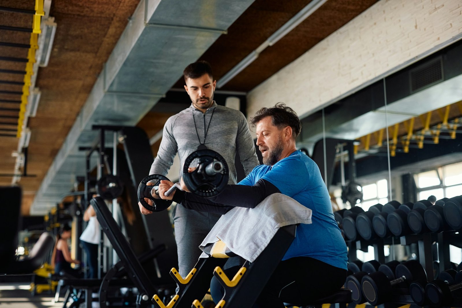 Mature man lifting weights while exercising with personal trainer in a gym.